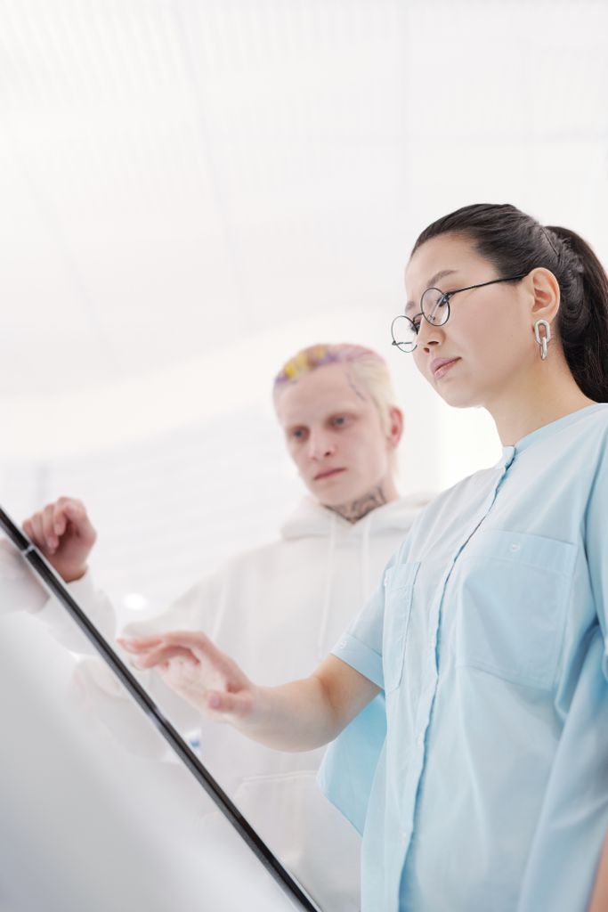 Asian woman with eyeglasses using an interactive display indoors, accompanied by a friend.