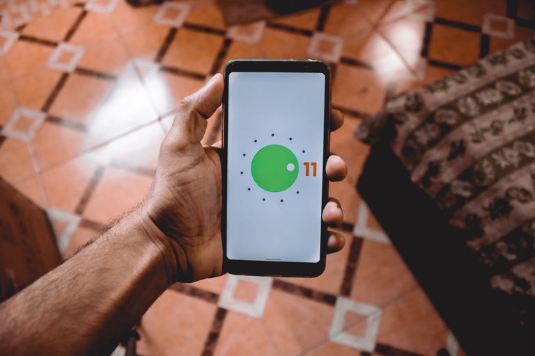 Pixel 11 AI Features: Close-up of a hand holding a smartphone displaying Android 11 interface indoors on patterned floor.