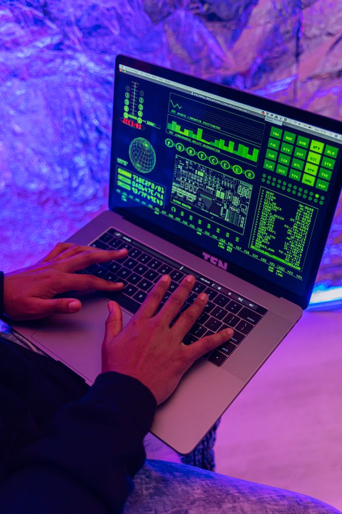 Close-up of hands typing on a laptop displaying green code, indoors, purple lighting.