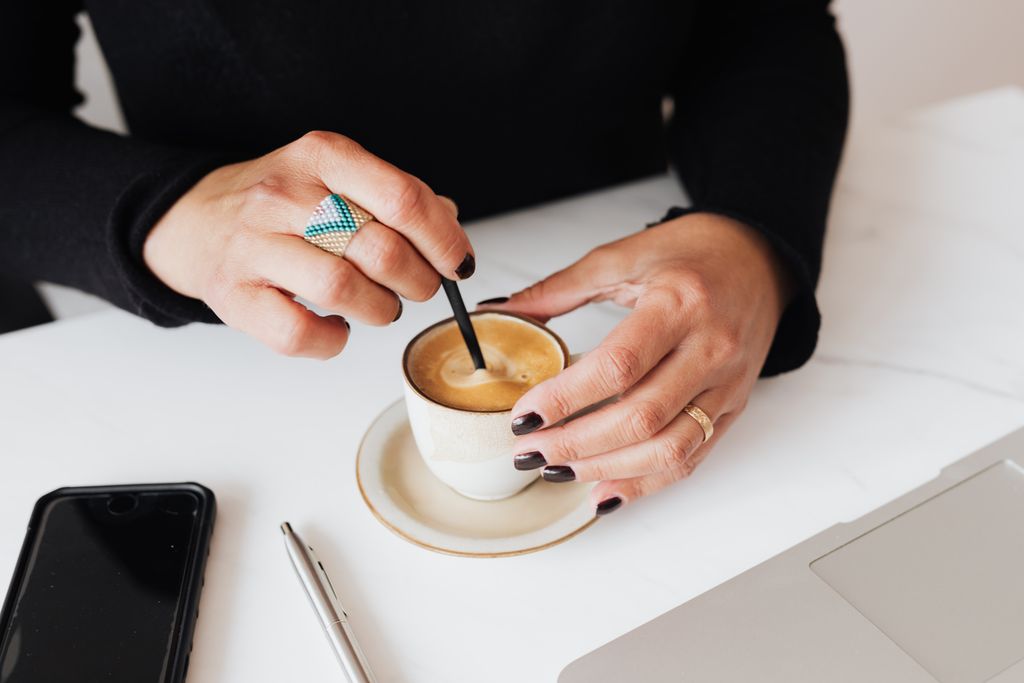 Crop unrecognizable female in black clothes sitting at table with laptop and smartphone while stirring cup of freshly brewed aromatic espresso
