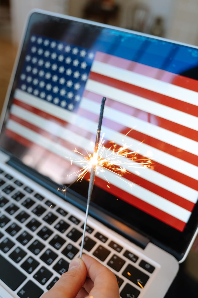Hand holding a sparker by a laptop displaying an American flag, symbolizing patriotic celebration.
