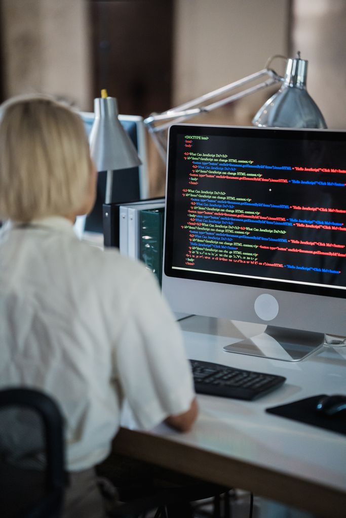 Professional woman coding at office desk with two monitors and desk lamps. Software Development