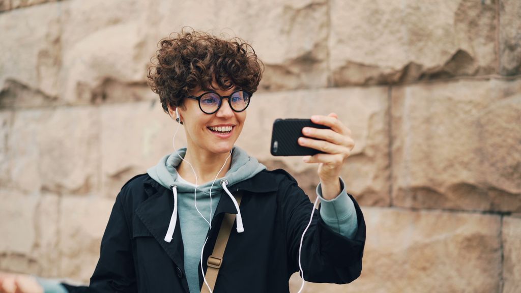 Smiling woman with curly hair taking a selfie in urban setting.