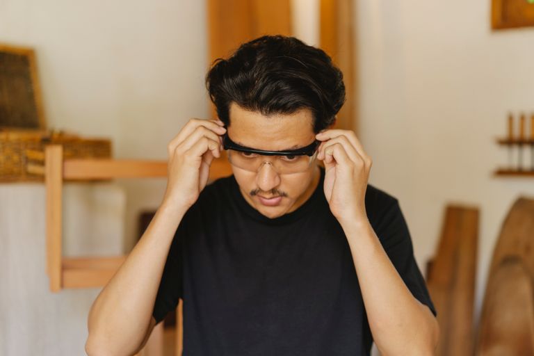 Young attentive ethnic male joiner in casual wear putting on carpentry glasses while looking down in workroom